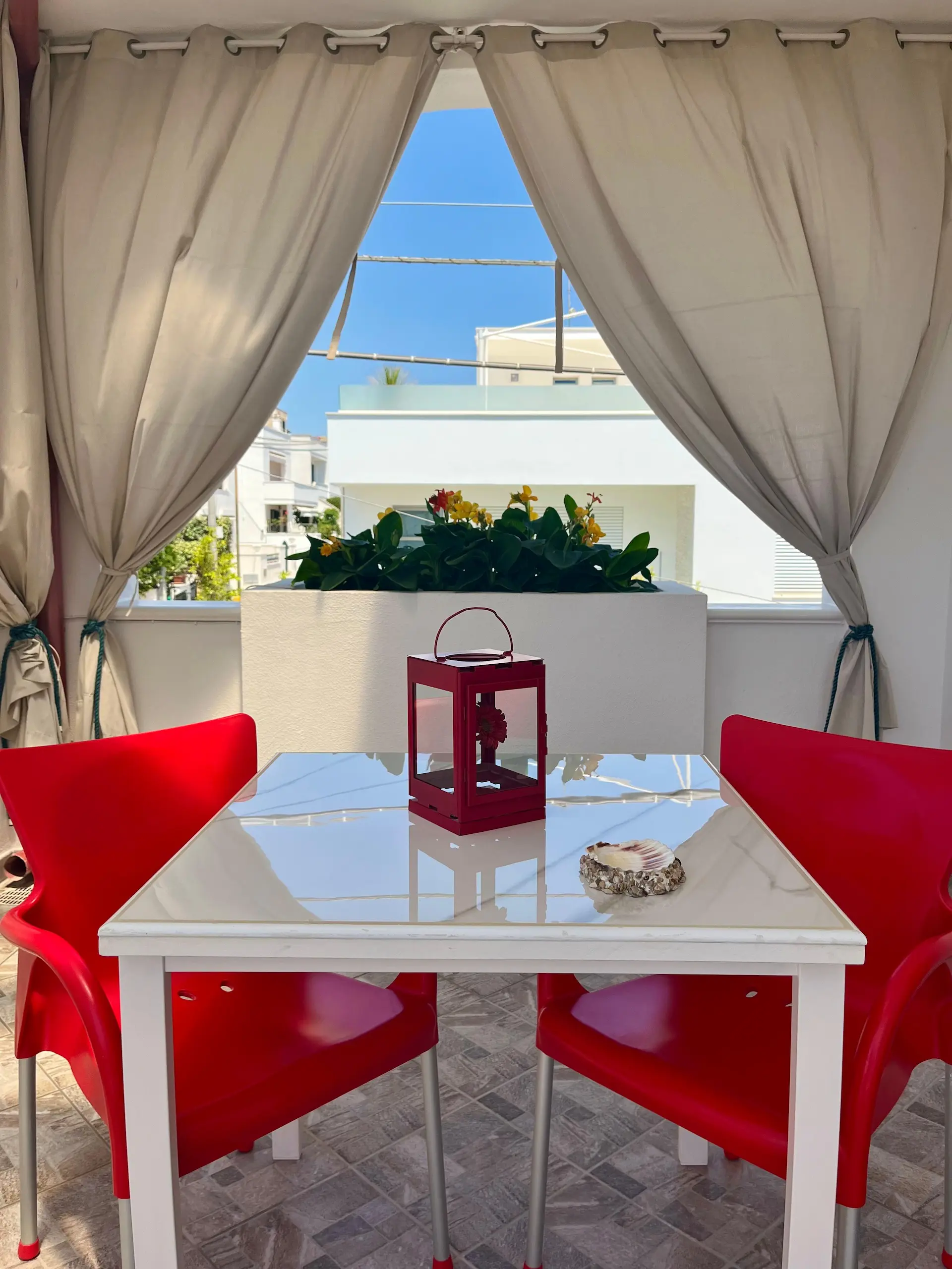 Veranda panoramica con fiori, tende beige, tavolo bianco e sedie rosse con vista mare e vista su Leuca - B&B Mareincantato